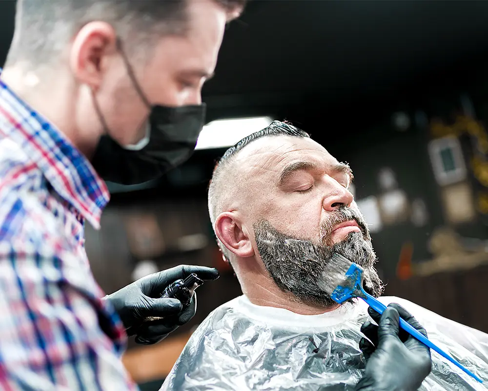 A man in a barbershop is having his grey beard hairs colored.